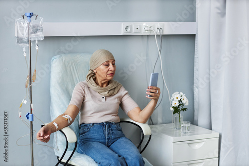 Photos Portrait of senior woman sitting in comfortable chair with IV drip and using sma