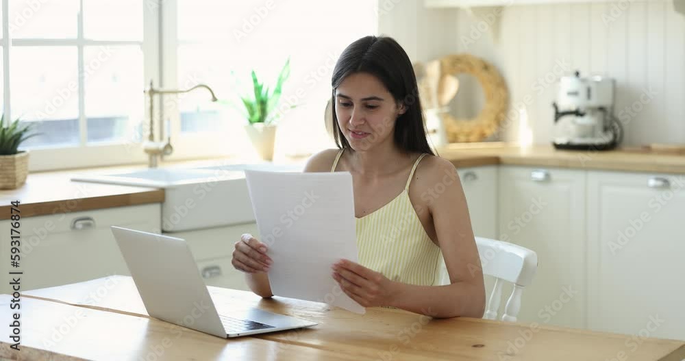 Focused engaged young adult girl reading paper document at laptop, getting good news, feeling happy, excited, throwing fist up in winner hand gesture, smiling, laughing, shouting yes, celebrating Focused engaged young adult girl reading paper document at laptop, getting good news, feeling happy, excited, throwing fist up in winner hand gesture, smiling, laughing, shouting yes, celebrating