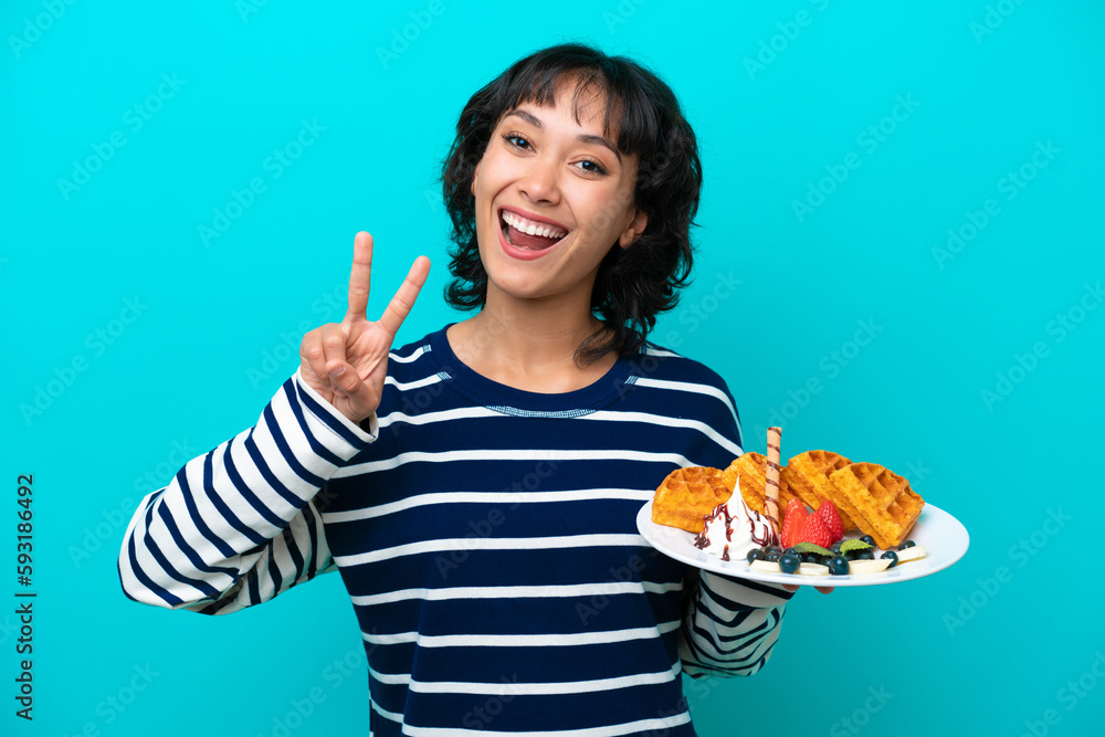 Young Argentinian woman holding waffles isolated on blue background smiling and showing victory sign Young Argentinian woman holding waffles isolated on blue background smiling and showing victory sign