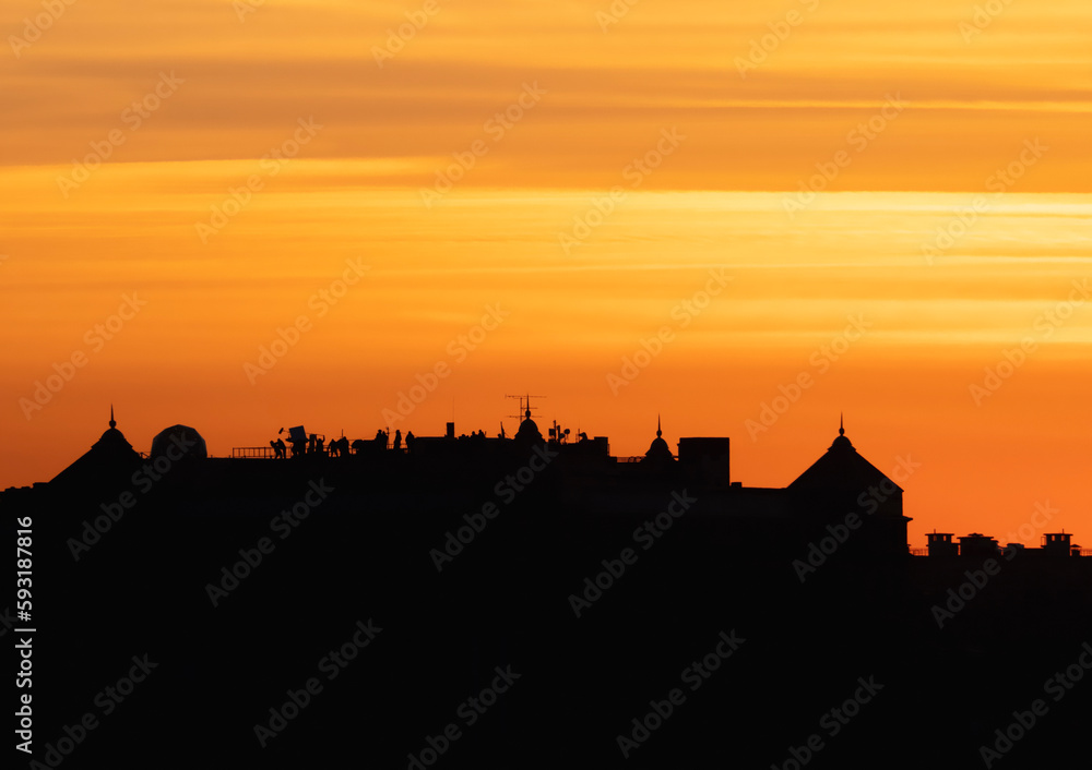 Contrasting view of dark silhouette of the roofs against the bright orange evening sky. Low angle view of long row of characteristic townhouses with roofs and chimneys of Europe houses during sunset