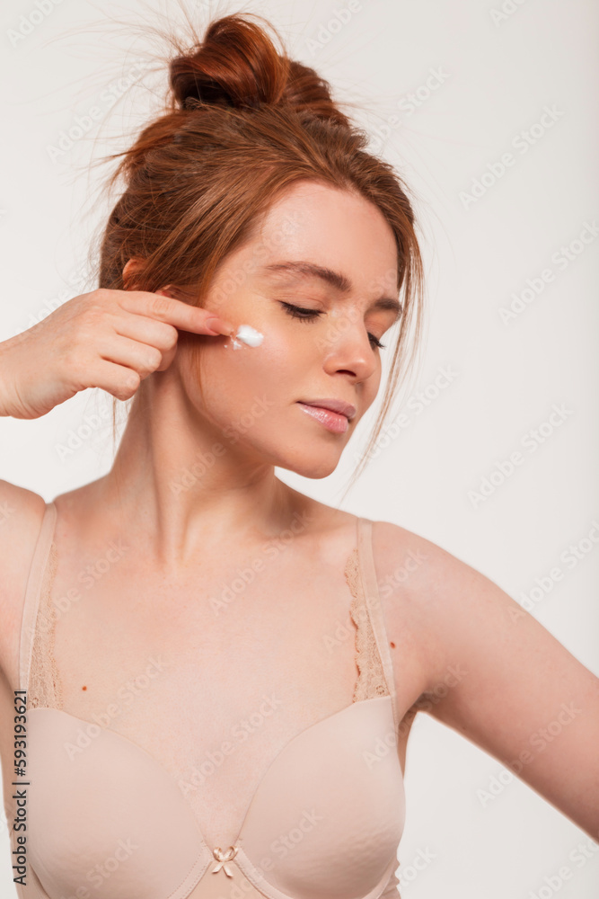 Beautiful young fresh redheaded woman with clear skin in a beige bra applies cream to face in the studio. Beauty and skin care Beautiful young fresh redheaded woman with clear skin in a beige bra applies cream to face in the studio. Beauty and skin care
