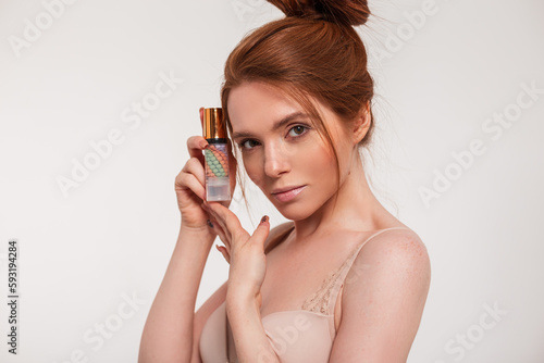 Beautiful red-haired young natural fresh girl holds cosmetics makeup base near her face and looks at the camera. Beauty, health and skin care
