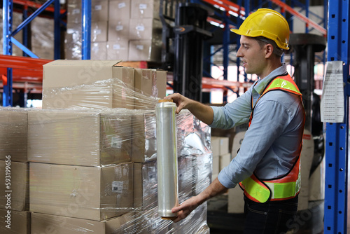 Caucasian warehouse worker man with hardhat and reflective jackets wrapping boxes in stretch film parcel on pallet while control stock and inventory in retail warehouse logistics distribution center