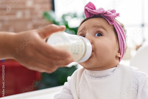 Adorable hispanic baby eating milk of feeding bottle sitting on highchair at home