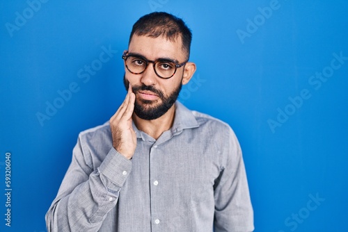 Middle east man with beard standing over blue background touching mouth with hand with painful expression because of toothache or dental illness on teeth. dentist