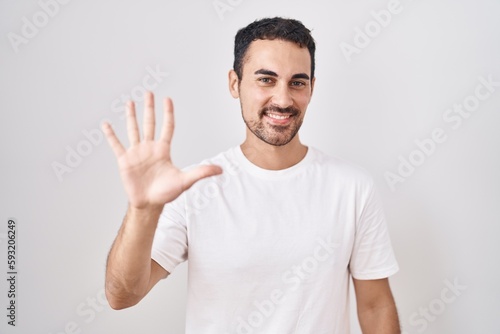 Handsome hispanic man standing over white background showing and pointing up with fingers number five while smiling confident and happy.