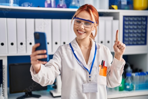 Young caucasian woman working at scientist laboratory doing video call with smartphone smiling with an idea or question pointing finger with happy face, number one