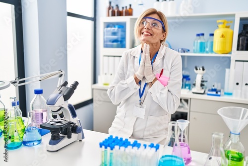 Middle age blonde woman working at scientist laboratory praying with hands together asking for forgiveness smiling confident.