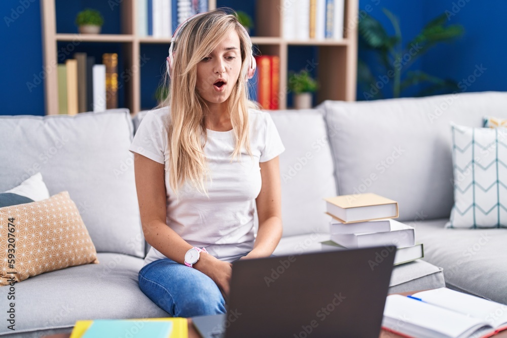 Young blonde woman studying using computer laptop at home afraid and shocked with surprise and amazed expression, fear and excited face.