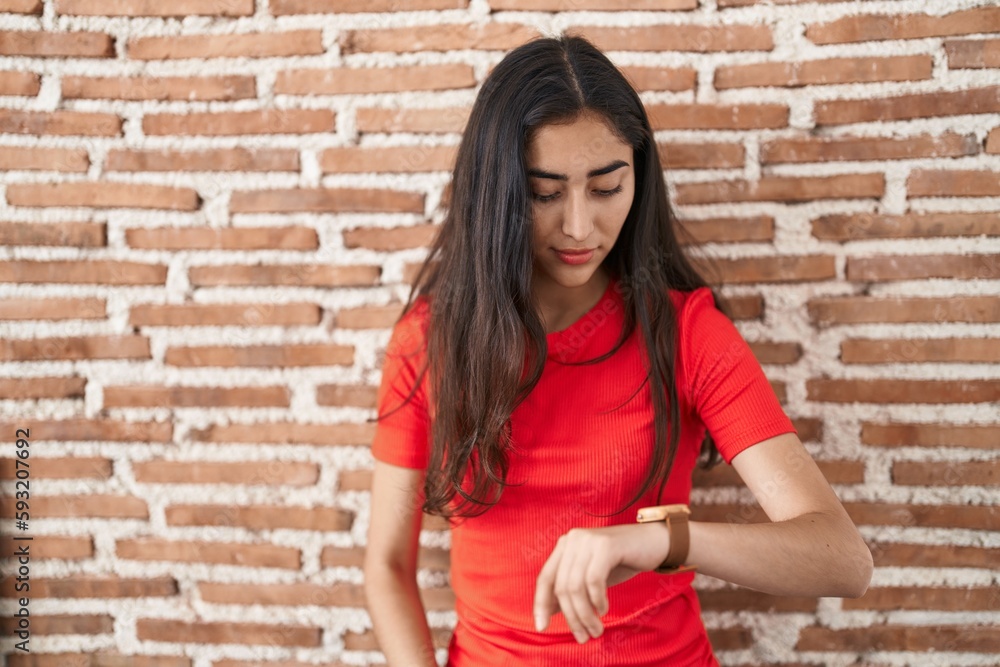 Young teenager girl standing over bricks wall checking the time on wrist watch, relaxed and confident Young teenager girl standing over bricks wall checking the time on wrist watch, relaxed and confident