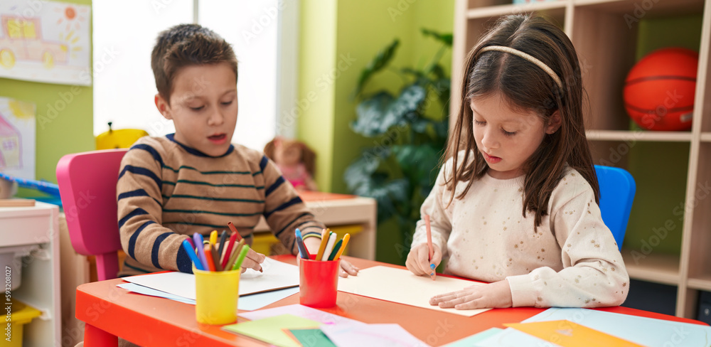 Fototapeta premium Adorable boy and girl preschool students sitting on table drawing on paper at kindergarten