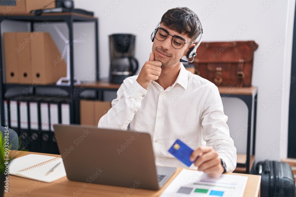 Young hispanic man working using computer laptop holding credit card with hand on chin thinking about question, pensive expression. smiling and thoughtful face. doubt concept. Young hispanic man working using computer laptop holding credit card with hand on chin thinking about question, pensive expression. smiling and thoughtful face. doubt concept.