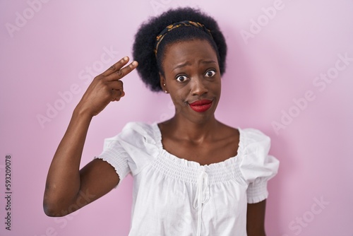 African woman with curly hair standing over pink background shooting and killing oneself pointing hand and fingers to head like gun, suicide gesture.