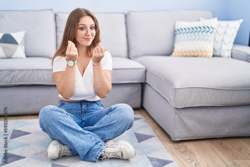 Young caucasian woman sitting on the floor at the living room doing money gesture with hands, asking for salary payment, millionaire business