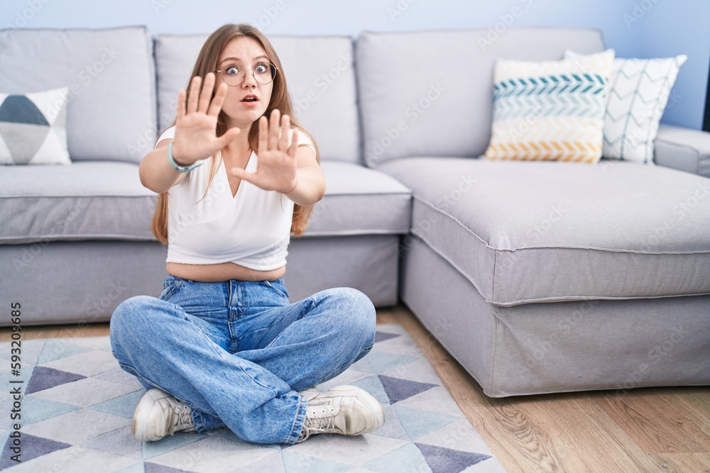 Young caucasian woman sitting on the floor at the living room afraid and terrified with fear expression stop gesture with hands, shouting in shock. panic concept. Young caucasian woman sitting on the floor at the living room afraid and terrified with fear expression stop gesture with hands, shouting in shock. panic concept.