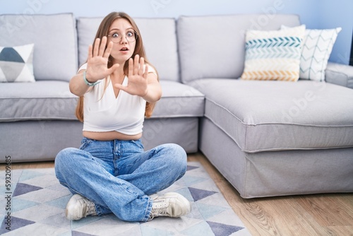 Young caucasian woman sitting on the floor at the living room afraid and terrified with fear expression stop gesture with hands, shouting in shock. panic concept.