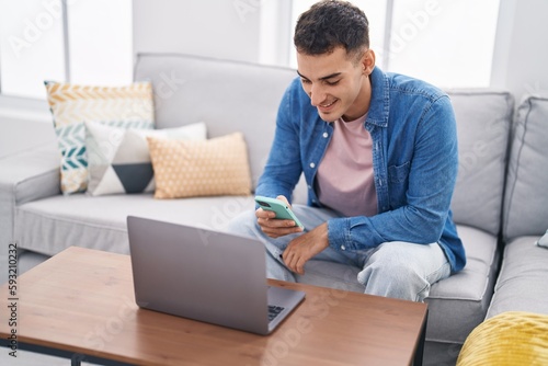 Young hispanic man using laptop and smartphone sitting on sofa at home