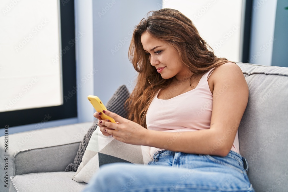 Young beautiful hispanic woman using smartphone sitting on sofa at home Young beautiful hispanic woman using smartphone sitting on sofa at home