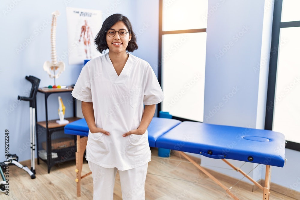 Young latin woman wearing physiotherapist uniform standing at physiotherapy clinic