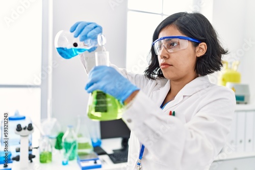 Young latin woman wearing scientist uniform pouring liquid on test tube at laboratory