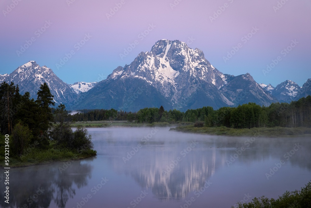 Purple sunset scene of snowy mountains range reflecting on a lake with trees