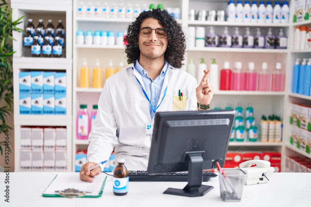 Hispanic man with curly hair working at pharmacy drugstore gesturing finger crossed smiling with hope and eyes closed. luck and superstitious concept. Hispanic man with curly hair working at pharmacy drugstore gesturing finger crossed smiling with hope and eyes closed. luck and superstitious concept.
