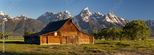 Panoramic view of an old wooden T.A. Moulton Barn in a green field against mountains in Wyoming