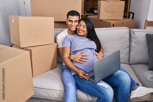 Young latin couple expecting baby touching belly using laptop at new home