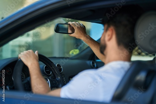 Young hispanic man driving car touching rearview at street