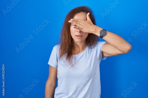 Brunette woman standing over blue background covering eyes with hand, looking serious and sad. sightless, hiding and rejection concept