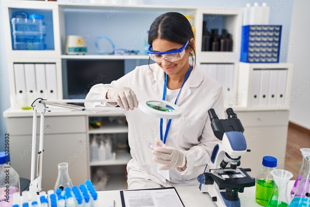 Young latin woman wearing scientist uniform using magnifying glass at laboratory Young latin woman wearing scientist uniform using magnifying glass at laboratory