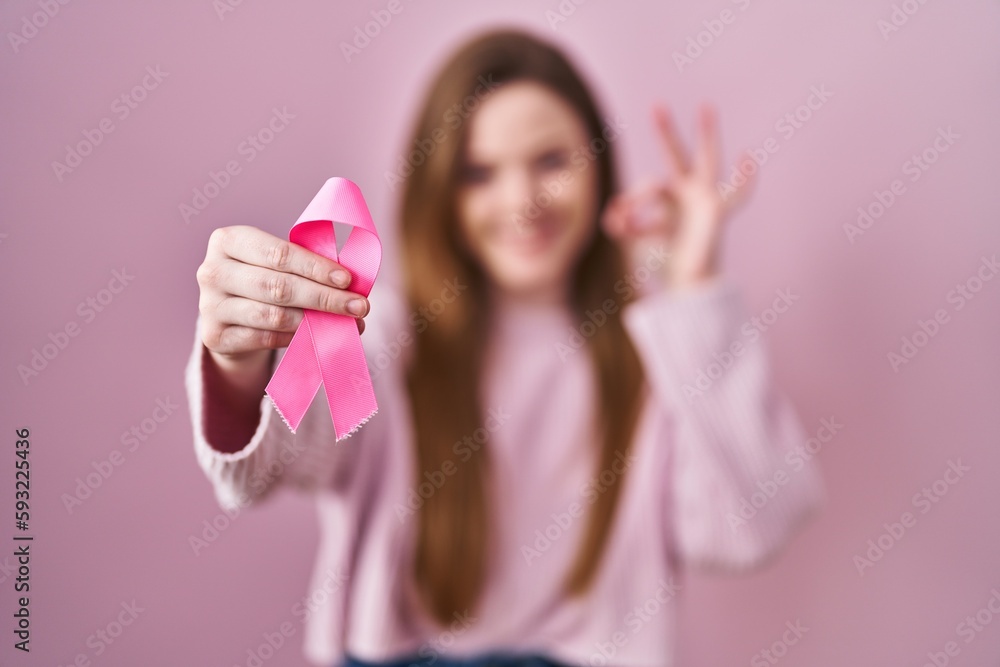 Young caucasian woman holding pink cancer ribbon doing ok sign with fingers, smiling friendly gesturing excellent symbol Young caucasian woman holding pink cancer ribbon doing ok sign with fingers, smiling friendly gesturing excellent symbol