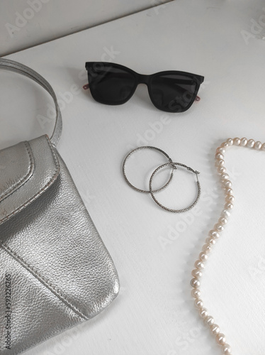 Photo flat lay of female fashion accessories, black sunglasses, silver purse, silver earrings, pearl necklace on a white table