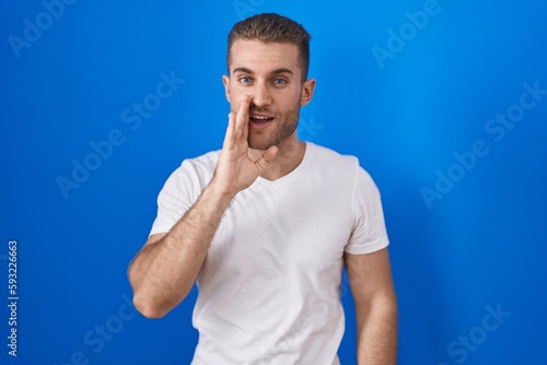 Young caucasian man standing over blue background hand on mouth telling secret rumor, whispering malicious talk conversation