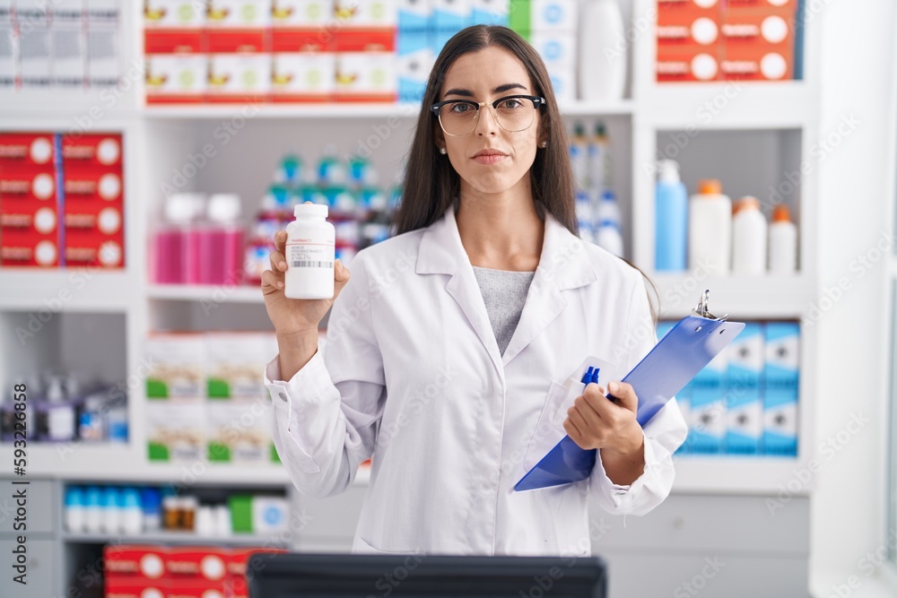 Young brunette woman working at pharmacy drugstore holding pills relaxed with serious expression on face. simple and natural looking at the camera. Young brunette woman working at pharmacy drugstore holding pills relaxed with serious expression on face. simple and natural looking at the camera.