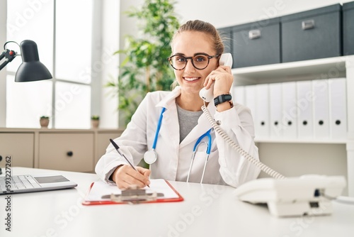 Young blonde woman wearing doctor uniform talking on the telephone working at clinic