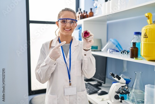 Young woman working at scientist laboratory holding geode smiling happy pointing with hand and finger
