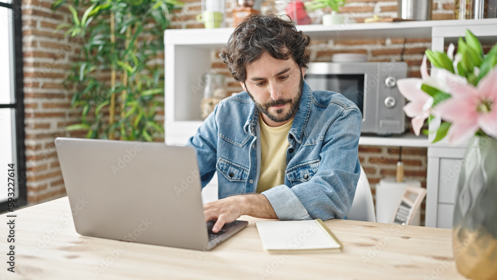 Young hispanic man using laptop reading notebook at dinning room Young hispanic man using laptop reading notebook at dinning room