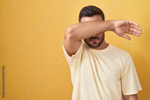 Handsome hispanic man standing over yellow background covering eyes with arm, looking serious and sad. sightless, hiding and rejection concept