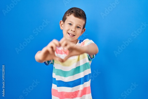 Young caucasian kid standing over blue background smiling in love doing heart symbol shape with hands. romantic concept.