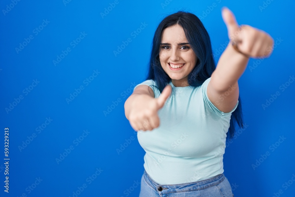 Young modern girl with blue hair standing over blue background approving doing positive gesture with hand, thumbs up smiling and happy for success. winner gesture. Young modern girl with blue hair standing over blue background approving doing positive gesture with hand, thumbs up smiling and happy for success. winner gesture.