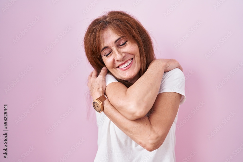 Middle age woman standing over pink background hugging oneself happy and positive, smiling confident. self love and self care Middle age woman standing over pink background hugging oneself happy and positive, smiling confident. self love and self care
