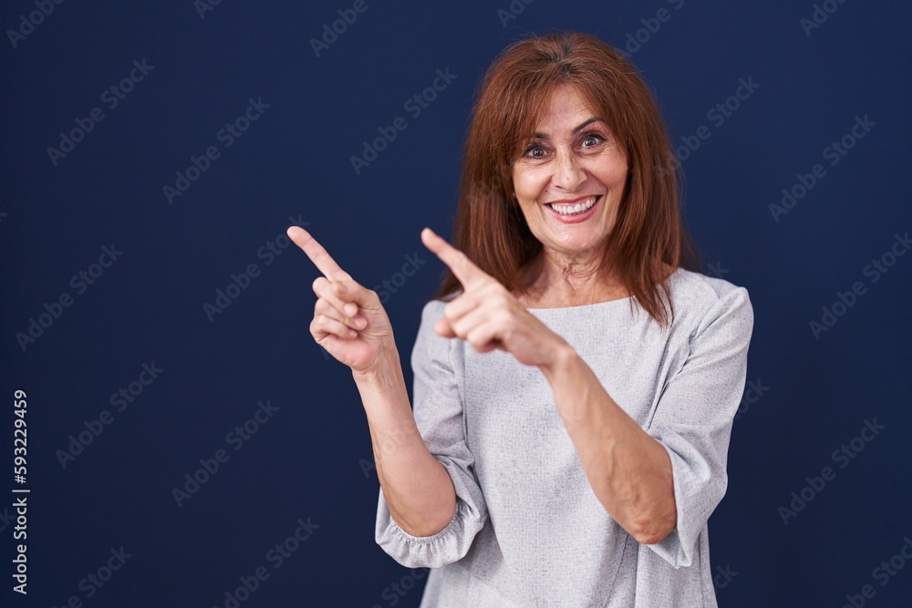 Middle age woman standing over blue background smiling and looking at the camera pointing with two hands and fingers to the side. Middle age woman standing over blue background smiling and looking at the camera pointing with two hands and fingers to the side.
