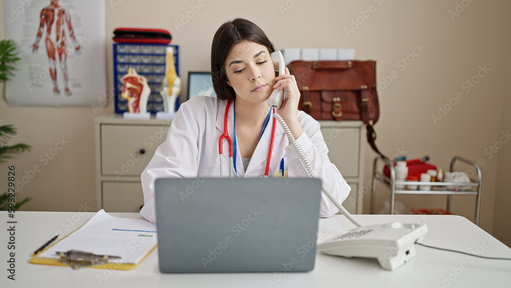 Young beautiful hispanic woman doctor using laptop talking on telephone at clinic