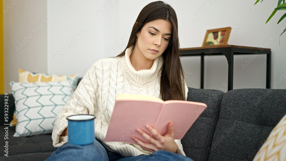 Young beautiful hispanic woman reading book and drinking coffee sitting on sofa at home Young beautiful hispanic woman reading book and drinking coffee sitting on sofa at home