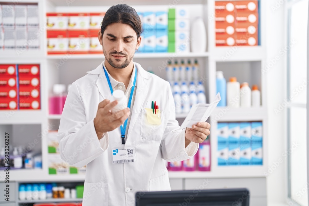 Young hispanic man pharmacist holding pills bottle reading prescription at pharmacy