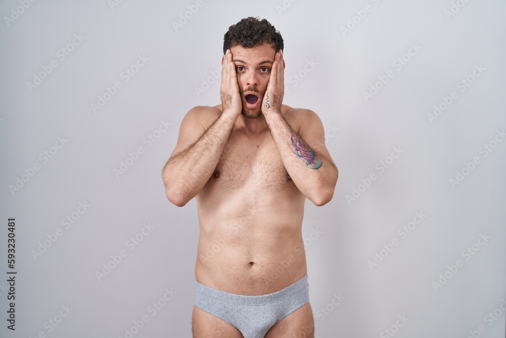 Young hispanic man standing shirtless wearing underware afraid and shocked, surprise and amazed expression with hands on face