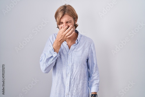 Caucasian man with mustache standing over white background smelling something stinky and disgusting, intolerable smell, holding breath with fingers on nose. bad smell