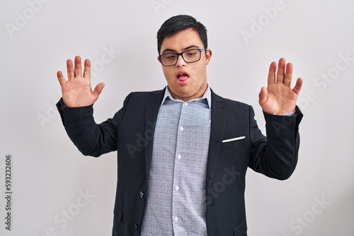 Young hispanic man with down syndrome wearing business style disgusted expression, displeased and fearful doing disgust face because aversion reaction. with hands raised