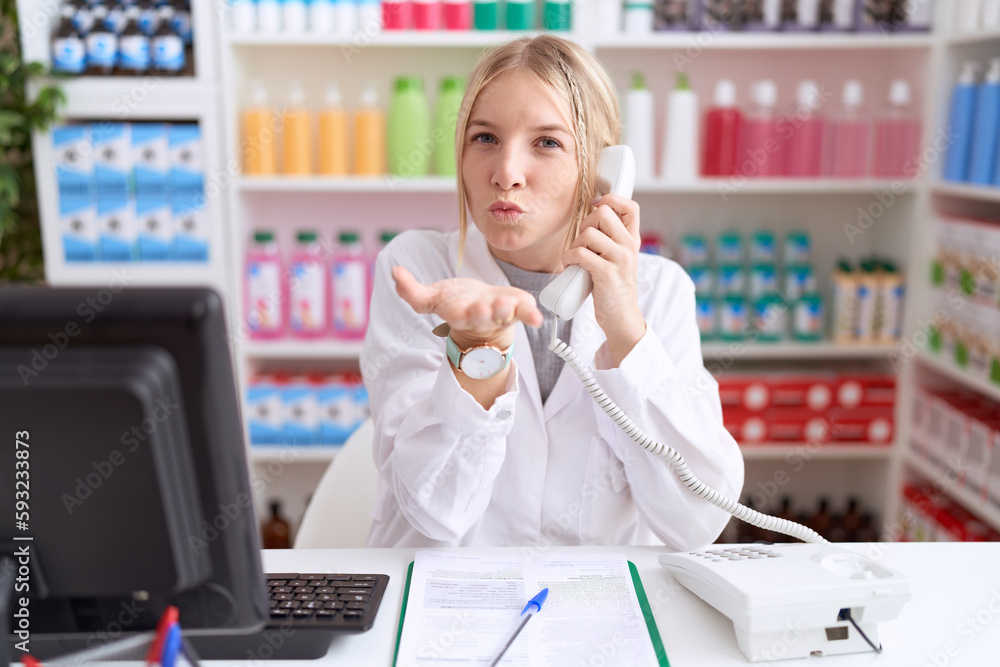 Young caucasian woman working at pharmacy drugstore speaking on the telephone looking at the camera blowing a kiss with hand on air being lovely and sexy. love expression.